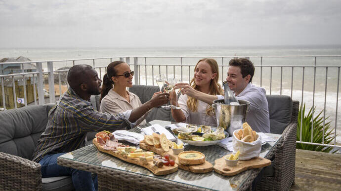 Two men and two women laugh together while sat on a hotel balcony with the sea beyond