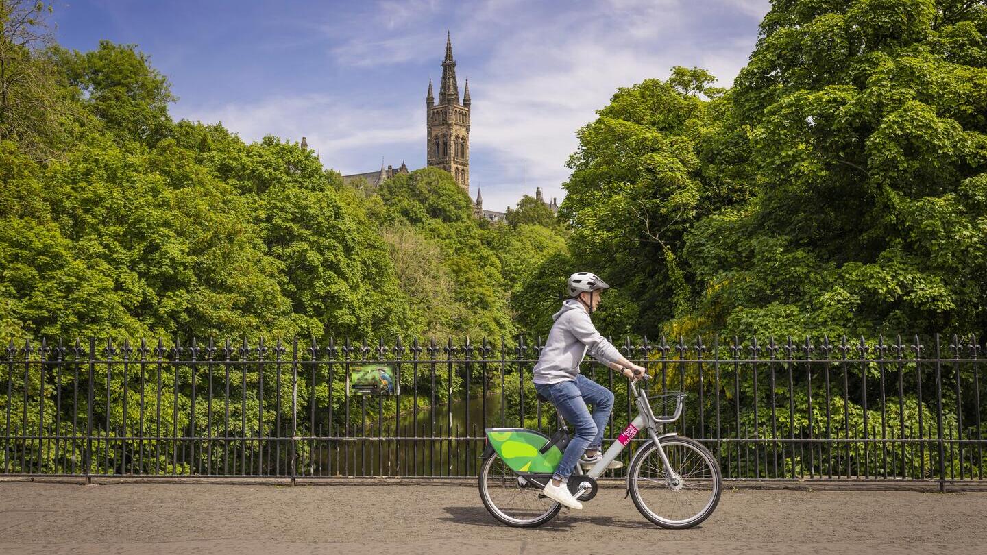 A Young Person Cycling An Ovo E-bike Over The River Kelvin in Glasgow’s popular Westend with a university and park in the background.
