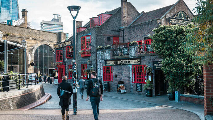 Street scene featuring The Anchor pub with red windows, historic brick buildings, modern skyscraper in background, and people walking.