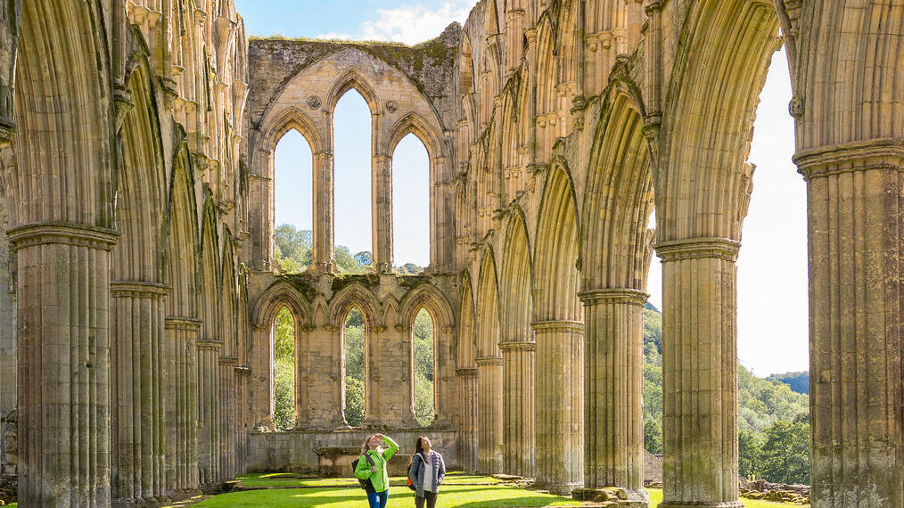 Two people walking between outdoor stone pillars of a ruined building