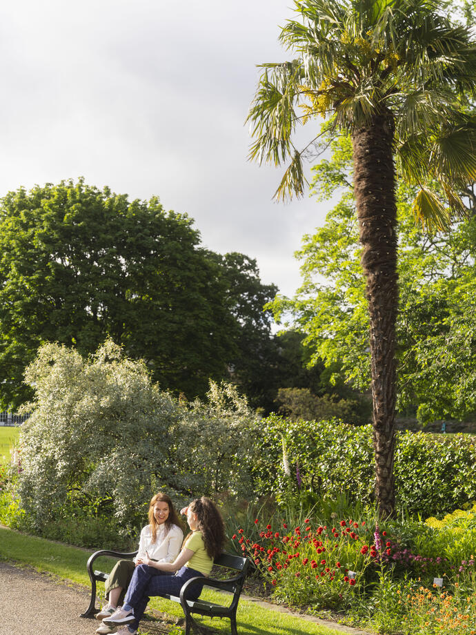 Two women sit on a bench in public gardens