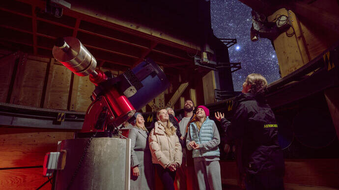 A group of people with an astronomer in an observatory, looking at the starry night sky beside a large telescope.