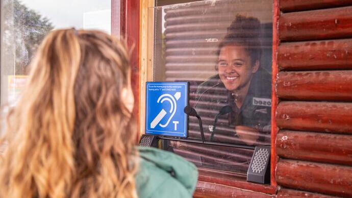 A person inside a ticket booth smiles at someone standing outside. A blue hearing loop sign is prominently displayed. Noahs Ark Zoo Farm - Gold award winner for the Accessible and Inclusive Tourism Award at the VisitEngland Awards for Excellence 2023.