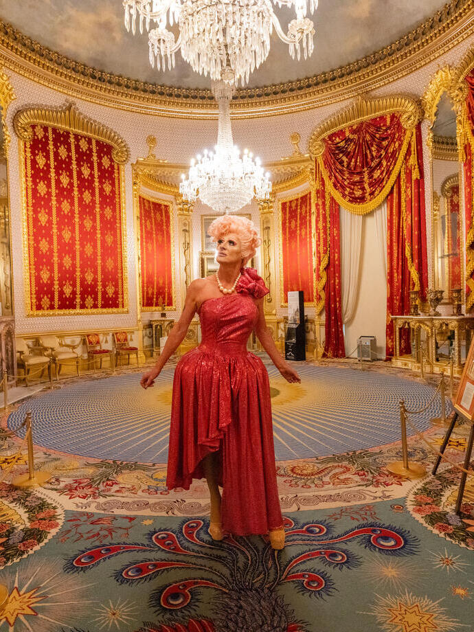 A drag performer wears a dress and stands in the lavish hall 
