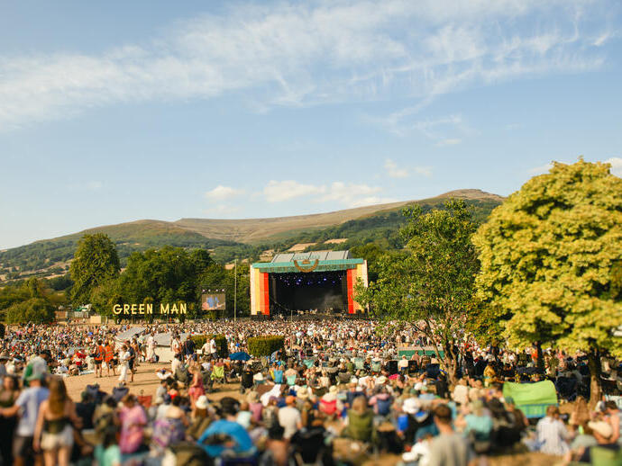 Large outdoor music festival crowd watching a stage, with mountains and trees in the background under a blue sky.