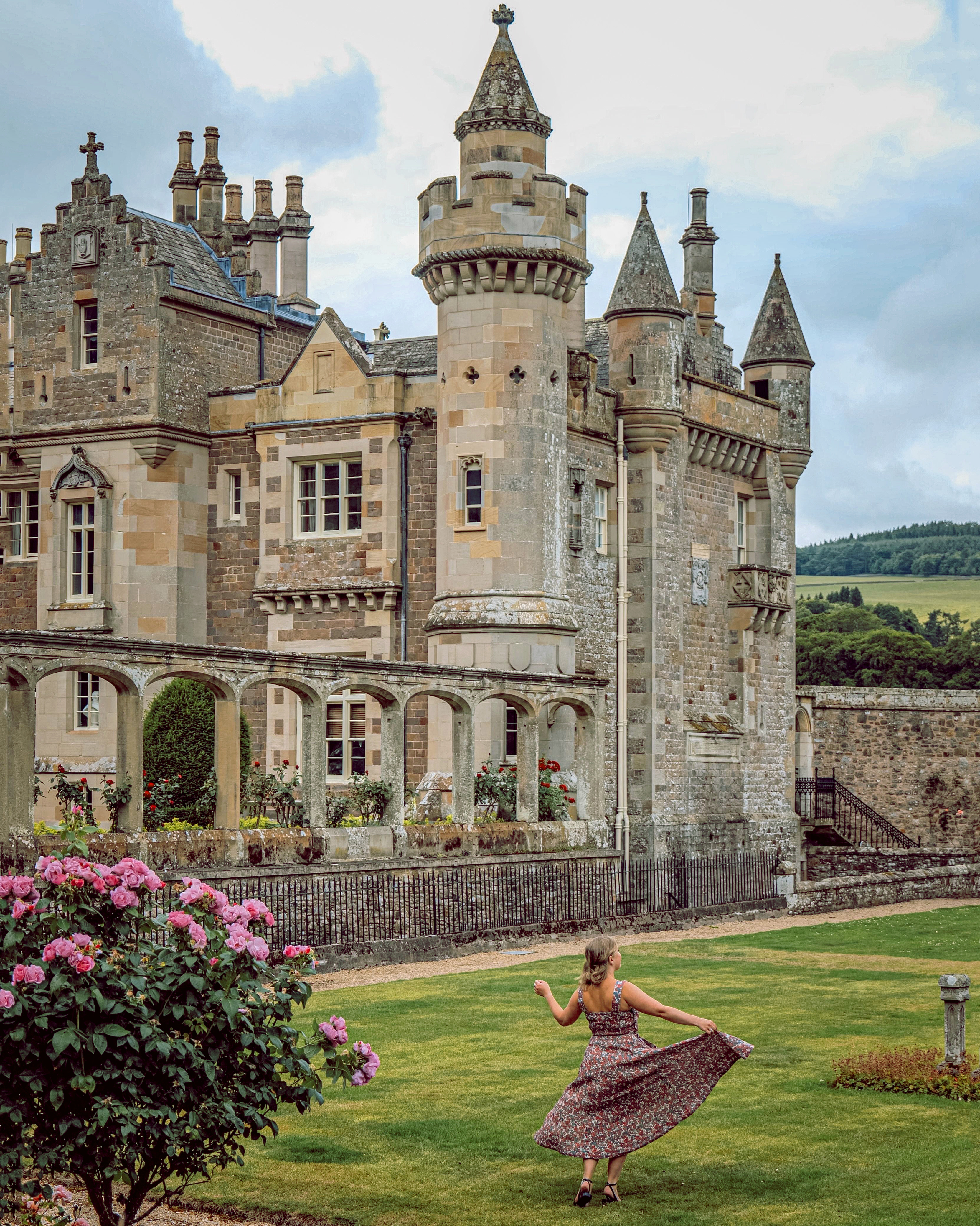 Historic stone castle with turrets and arched walkway, lush gardens, pink flowers, and a woman twirling on the lawn under a cloudy sky.