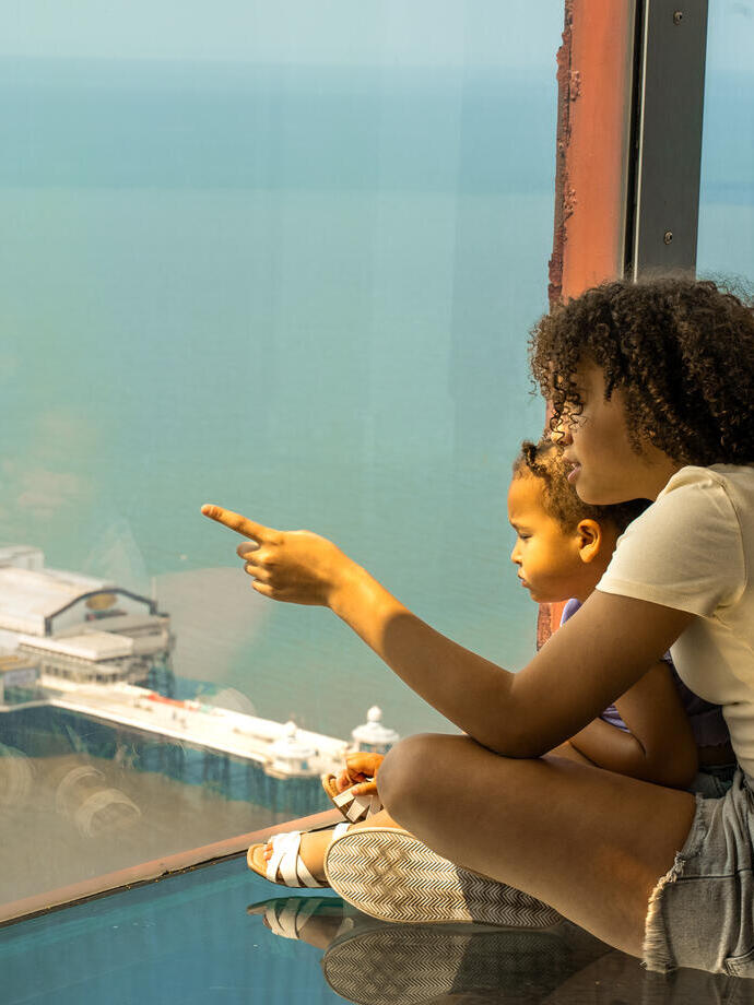 Two people sit by a large window overlooking the sea, with a pier and building visible below. One person points at the view.