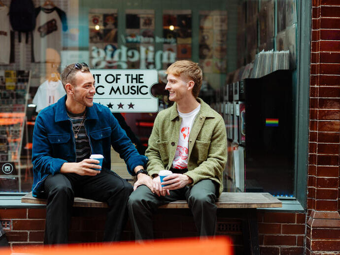 Two men sit with take away drinks in front of a shop
