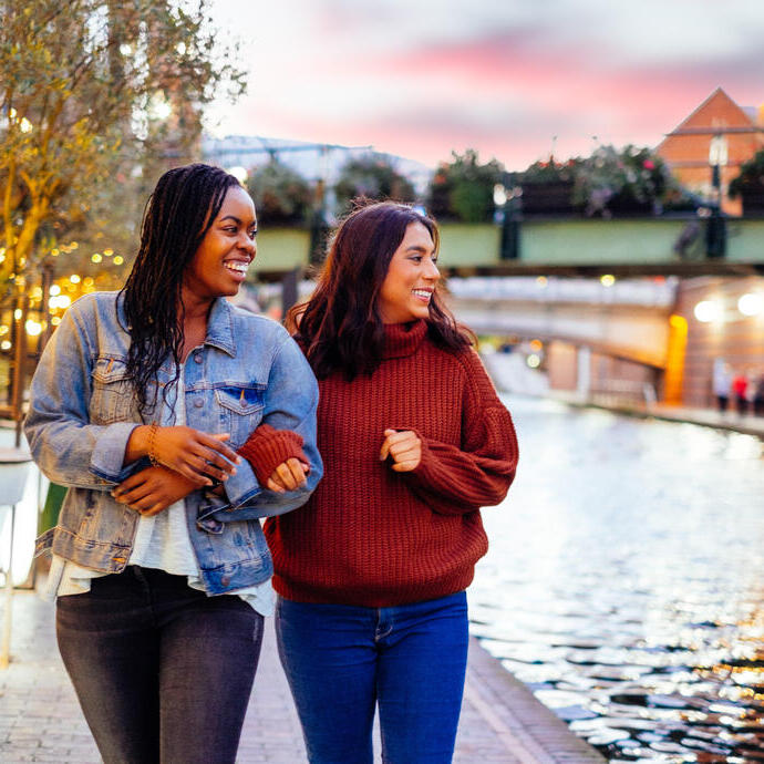 Two women, linking arms, walking beside a canal in evening