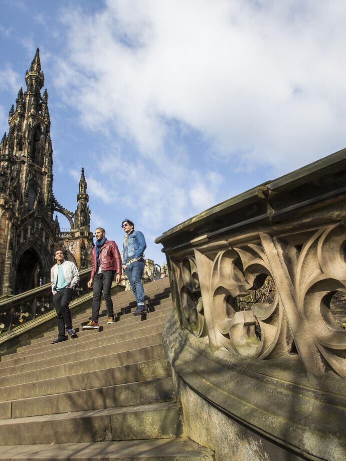 Three people walking down stone steps near the ornate Scott Monument under a partly cloudy blue sky in Edinburgh.