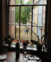 View through a window onto a cobblestone street with two people walking, surrounded by historic brick and pastel buildings.