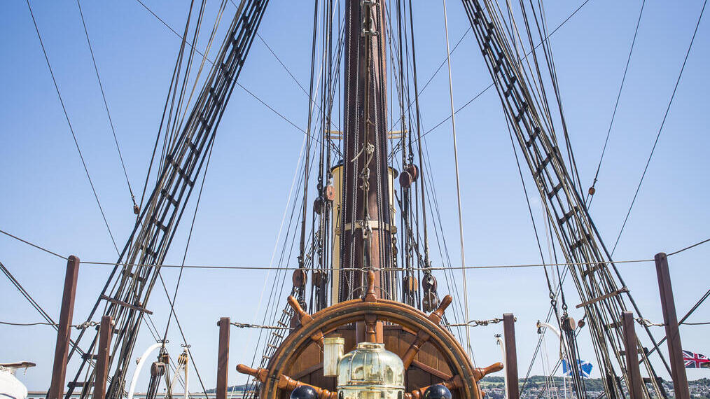 Wooden ship's wheel and rigging on the deck of a historic sailing vessel, with blue sky and seaside background.