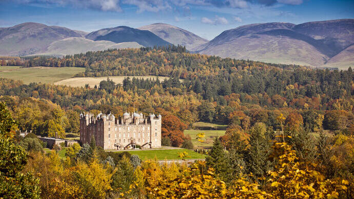 A historic castle set amid vibrant autumn trees, green fields, and hills under a bright blue sky.