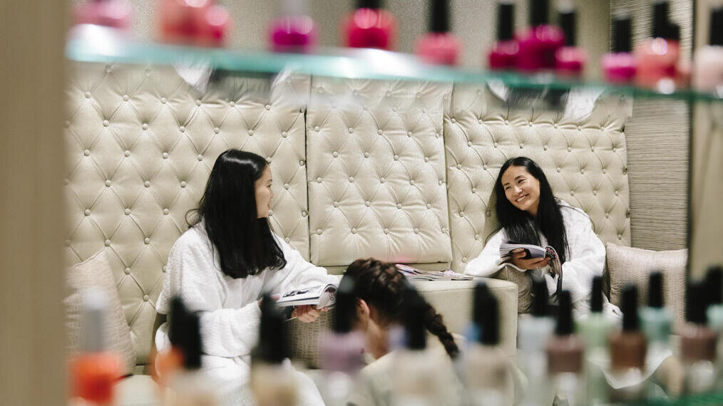 Two women, sat in white robes in a spa reading magazines