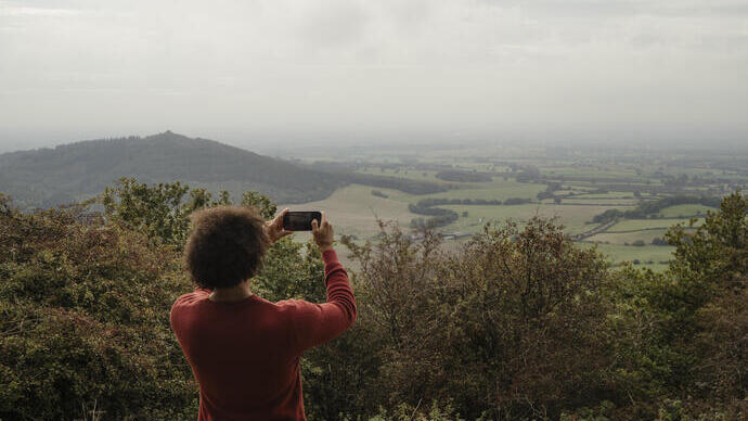 Person in a red sweater taking a photo of a scenic landscape with rolling hills, fields, and cloudy sky from a viewpoint.