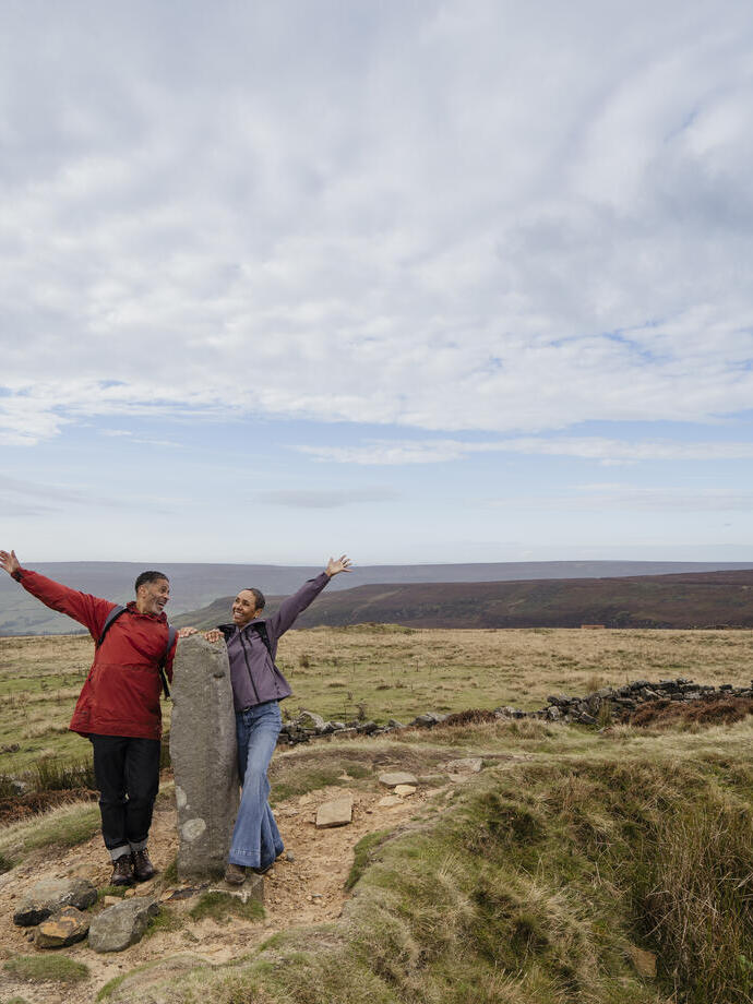 Two people posing cheerfully beside a standing stone in open countryside under a wide, partly cloudy sky. Rolling hills and grassy moorland in the background.