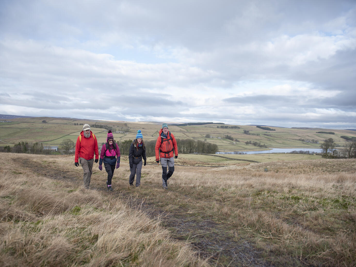 Four people hiking on grassy hills with patchwork fields and a lake in the background under a cloudy sky.