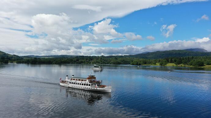 Cruise boats sail on a calm lake surrounded by green hills and forest under a partly cloudy blue sky.