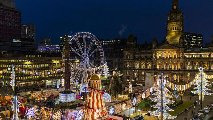 A festive night scene featuring a brightly lit Ferris wheel, holiday market, illuminated decorations, and a historic building in the background.