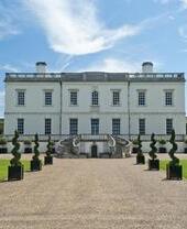Gran mansión blanca con jardín, entrada adornada con árboles podados y cielo azul con nubes.