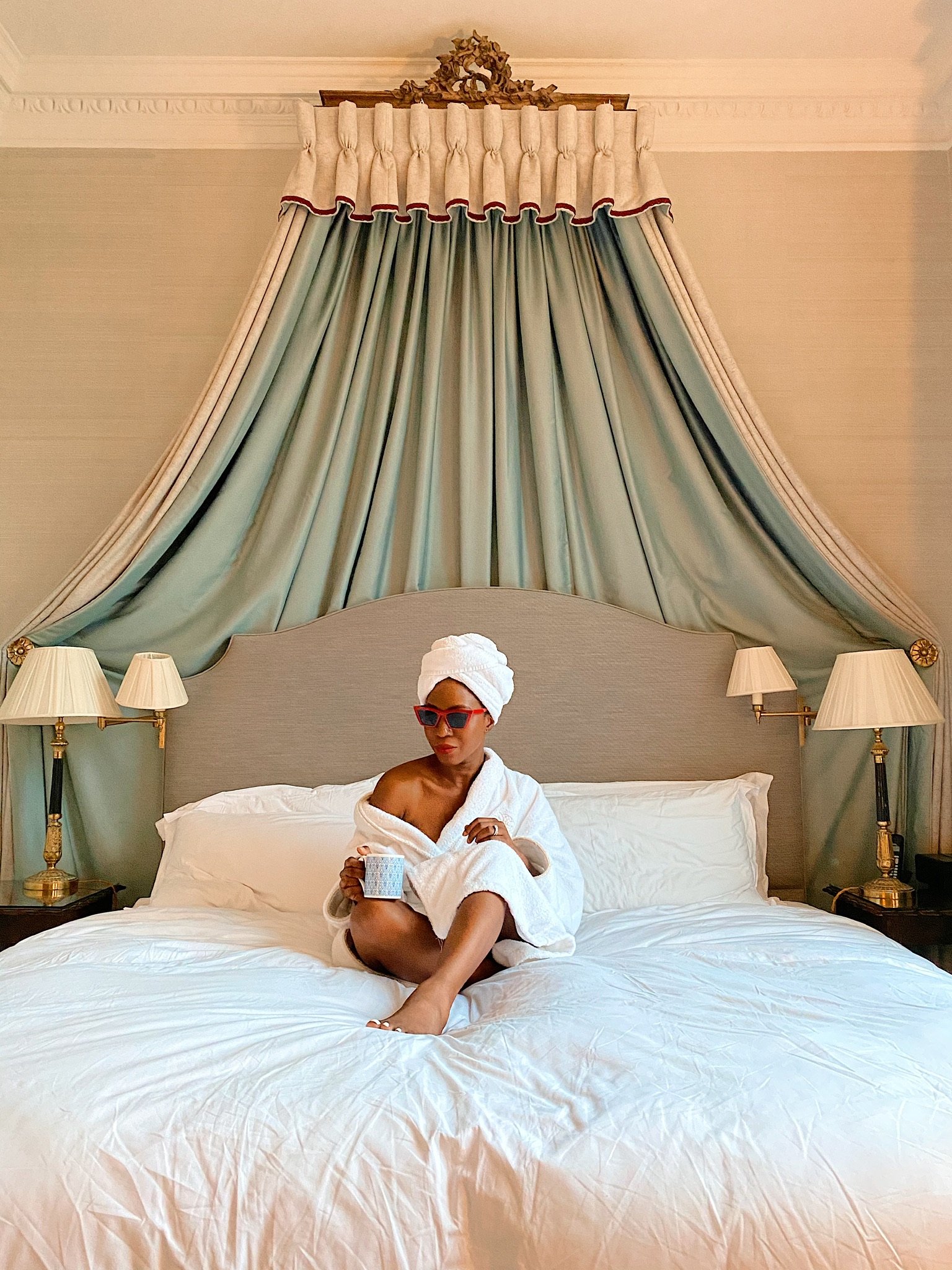 Woman sits on a bed wearing a robe and sunglasses in a hotel bedroom