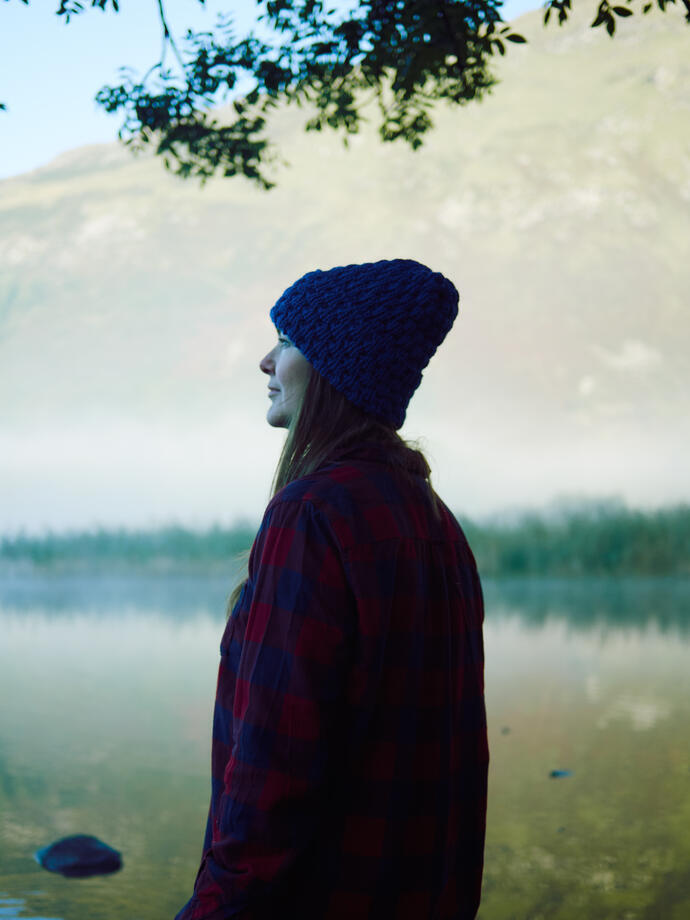 Femme coiffée d'un chapeau debout devant un lac avec de la brume et des montagnes en arrière-plan.