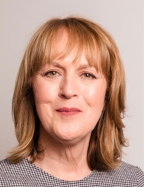 Portrait of a middle-aged woman with shoulder-length light brown hair, wearing a black and white patterned top, smiling slightly, with a plain background.