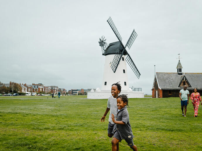 Children run on a green field near a traditional white windmill and buildings, with adults walking in the background on a cloudy day.