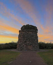 A small stone tower with a plaque on it surrounded by grass and moor under a vibrant blue and orange sky at sunset.