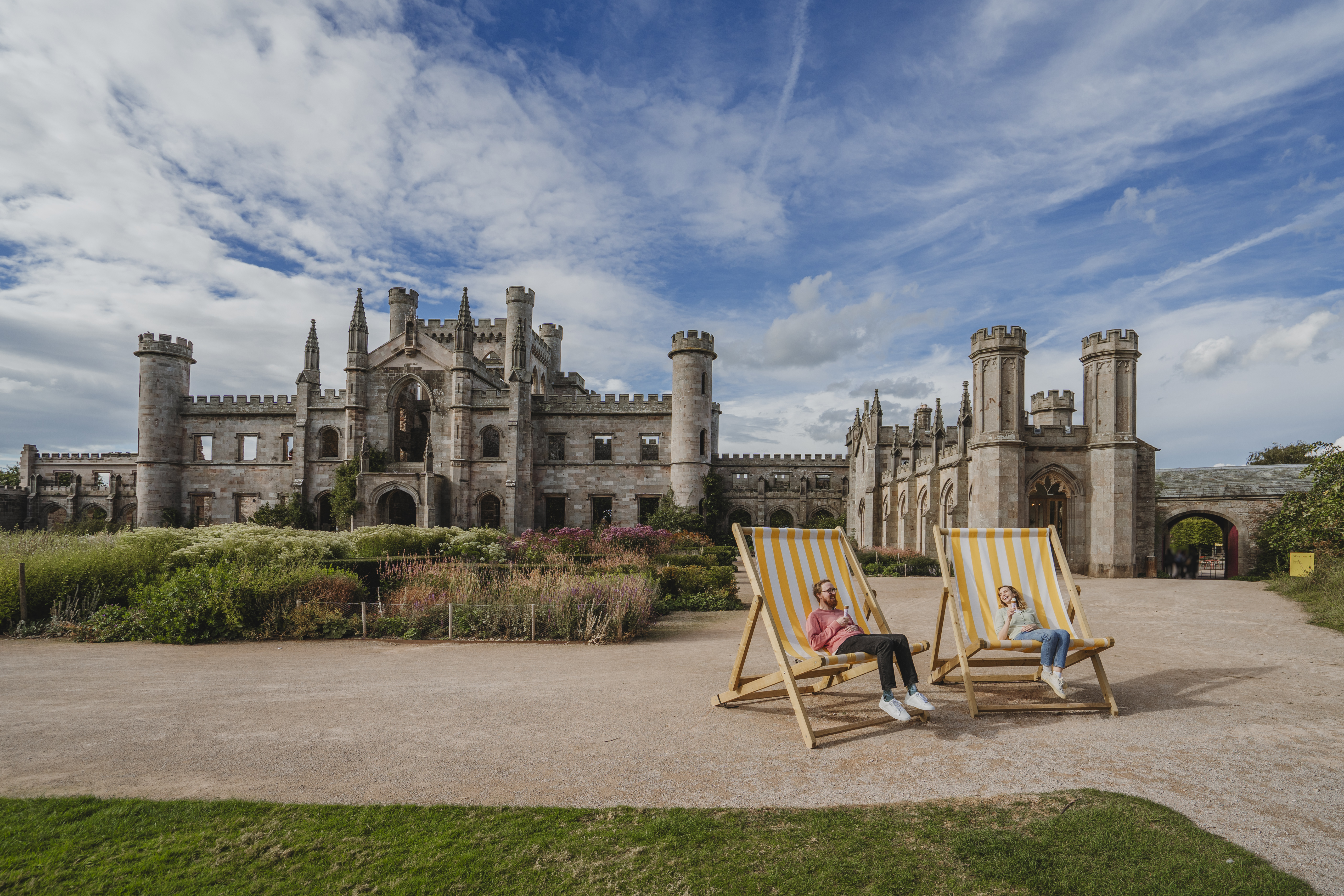 Zwei Personen sitzen auf großen Liegestühlen vor einem historischen britischen Schloss unter teilweise bewölktem Himmel.