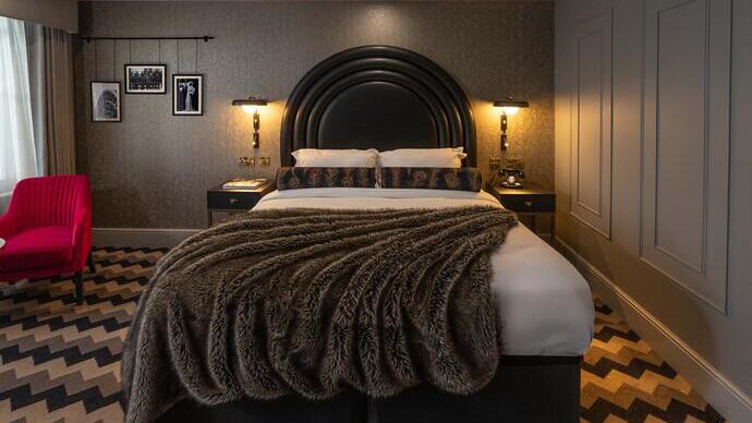 Modern hotel room with a large bed, dark curved headboard, patterned carpet, red chair, and stylish lighting. Monochrome wall art and neutral tones dominate the decor.