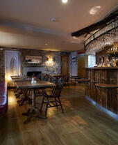 Interior of a pub with the bar area, showing bench seating and dining room tables and chairs, exposed stone walls and a stone fireplace in background