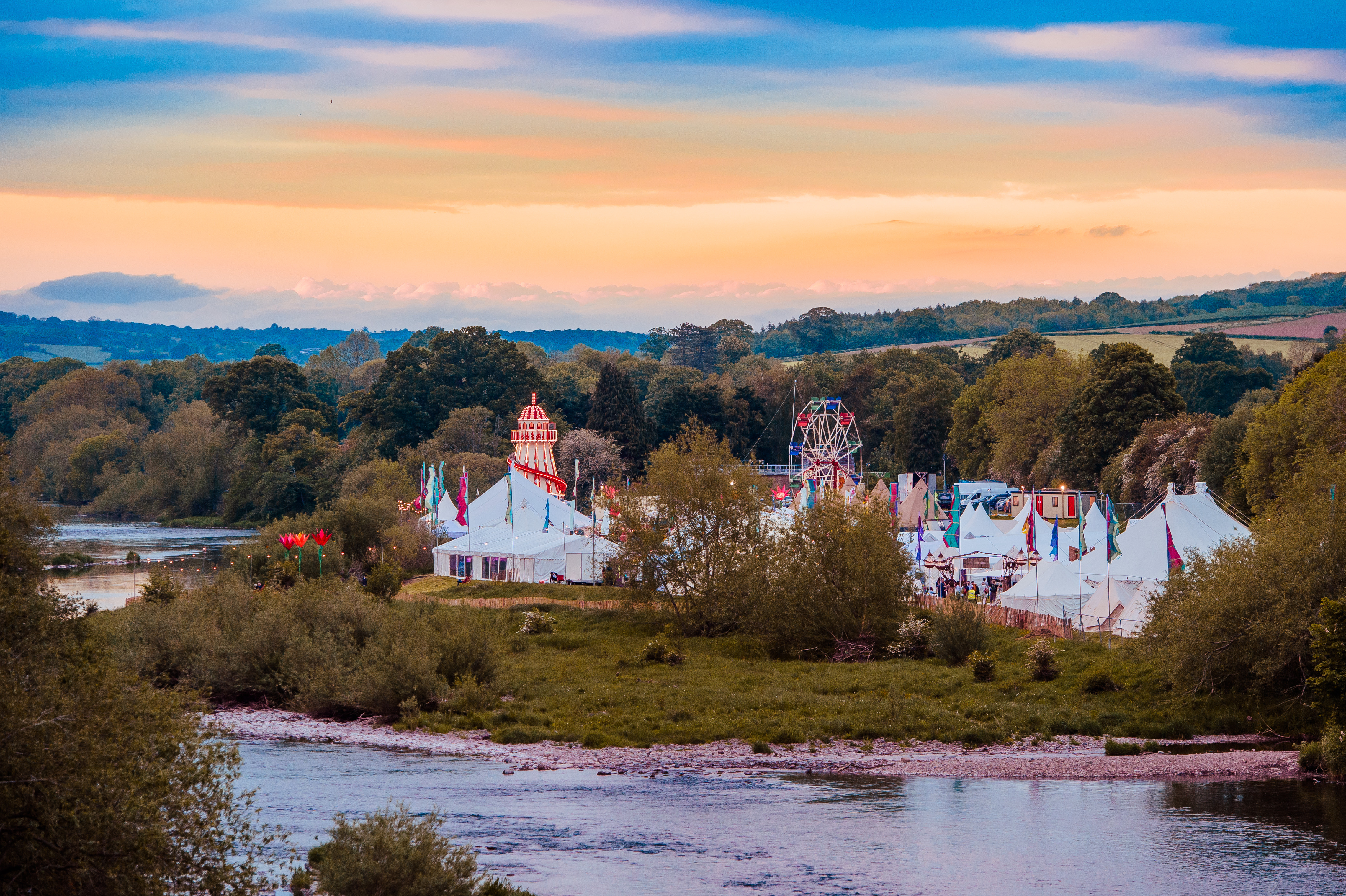 Tents and fairground rides of a summer festival beside a river.