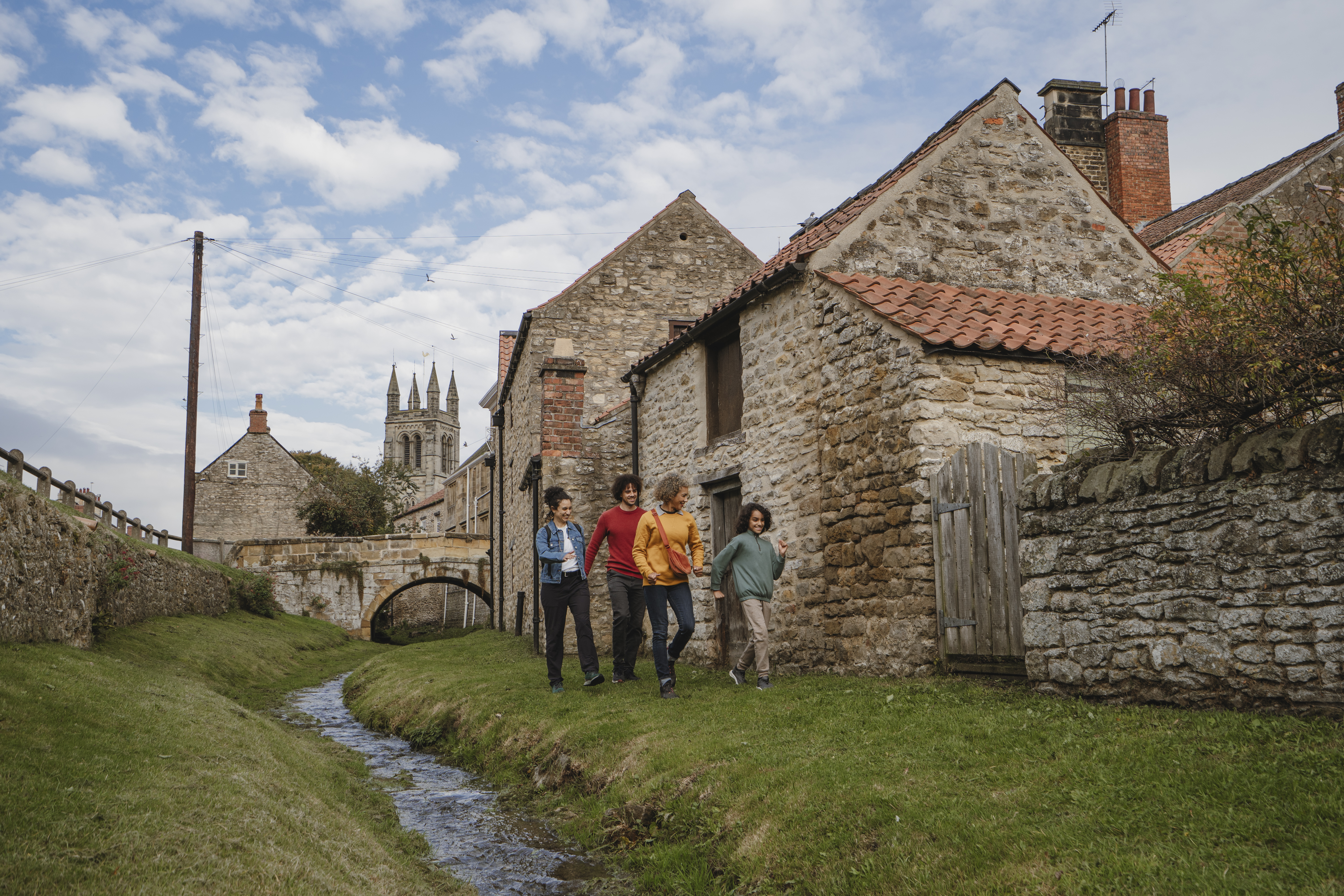 Four people walk beside a stone cottage and stream in a historic village, with a church tower and arched bridge in the background under a partly cloudy sky.