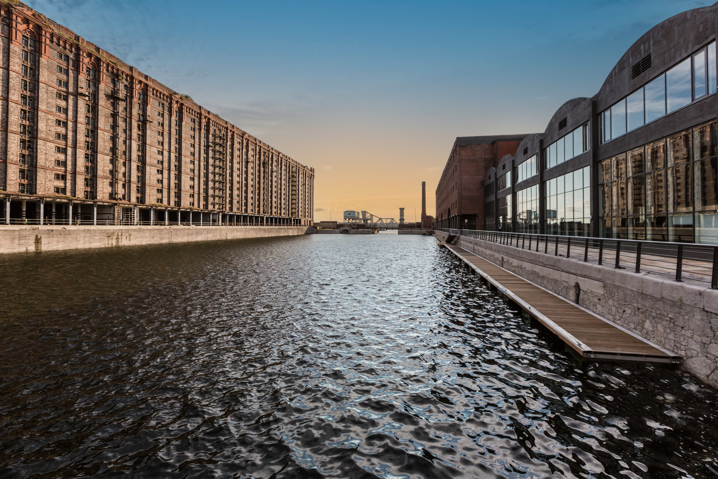 A view of a dock looking across the water. To the left stands a traditional brick built dockside building, to the right is a modern building made from glass and wood.