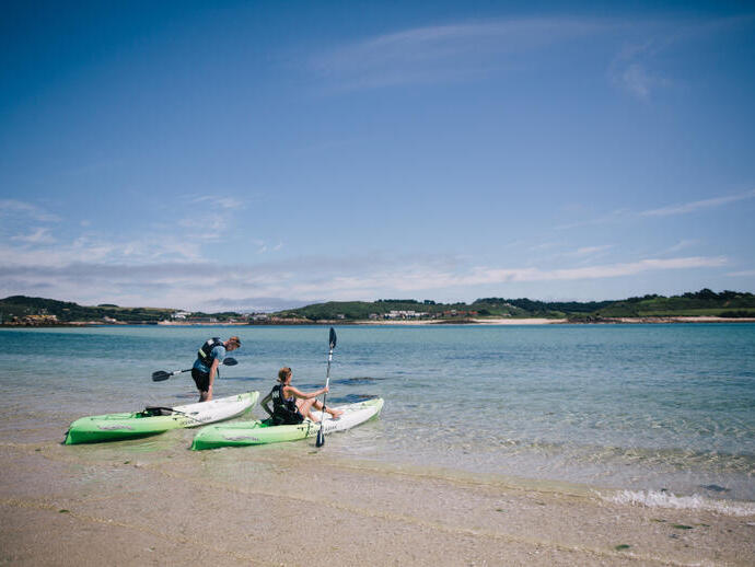 Two people with kayaks on a sandy shore beside clear blue water, with green hills in the background under a blue sky.