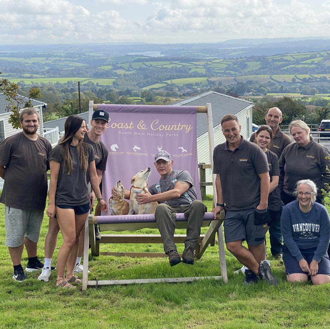 Group of people and a dog posing by a Coast & Country sign outdoors, with rolling countryside and lodges in the background.