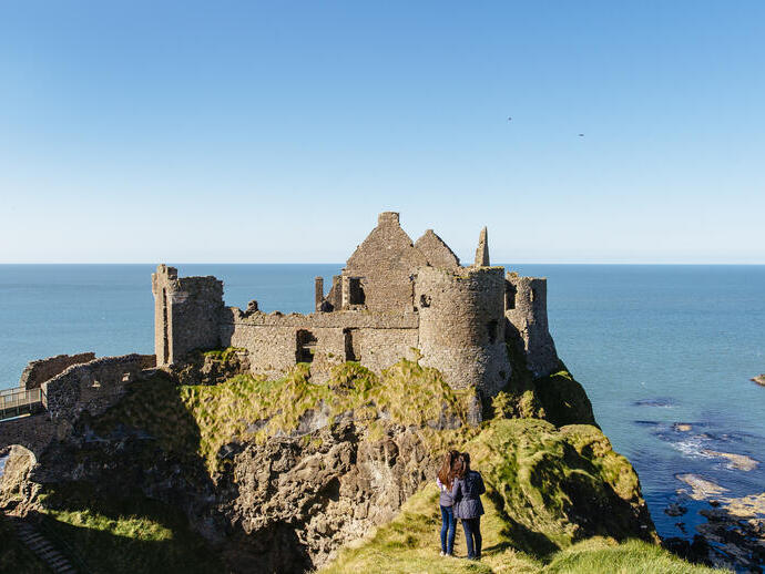 People looking at an historic stony castle from a rocky outcrop cliff over the ocean.