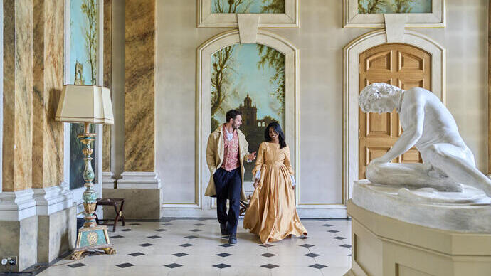 Elegant hall with classical decor, statue, ornate lamp, and two people in period attire walking and talking. Arched murals and wooden doors in the background.