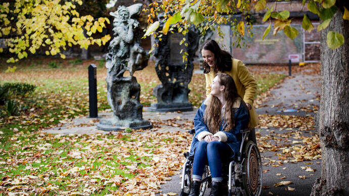 Two people, one in a wheelchair and one standing, enjoy a park with autumn leaves and abstract outdoor sculptures.