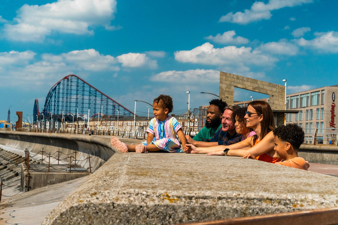 Family sits on seaside promenade with a rollercoaster and modern buildings in the background under a blue sky with clouds.