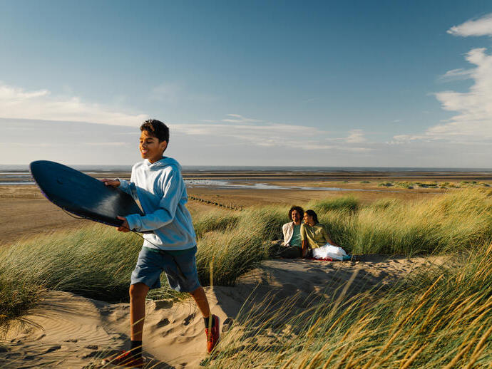 Boy with a surfboard walks on sandy dunes, while two people relax in long grass beside a wide, empty beach under a bright blue sky.