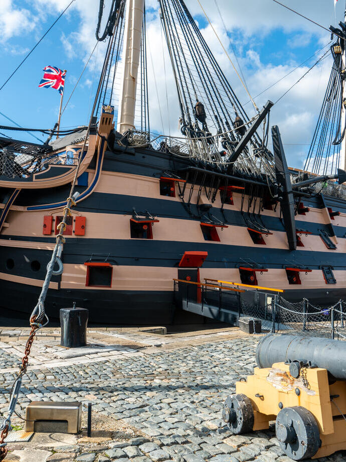 Historic wooden warship docked on cobblestone with a cannon in the foreground, under a blue sky with some clouds.