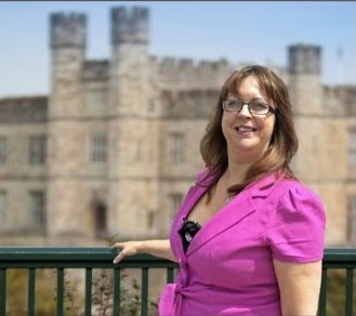 Person in a bright pink outfit standing in front of a historic stone castle with towers, on a sunny day.