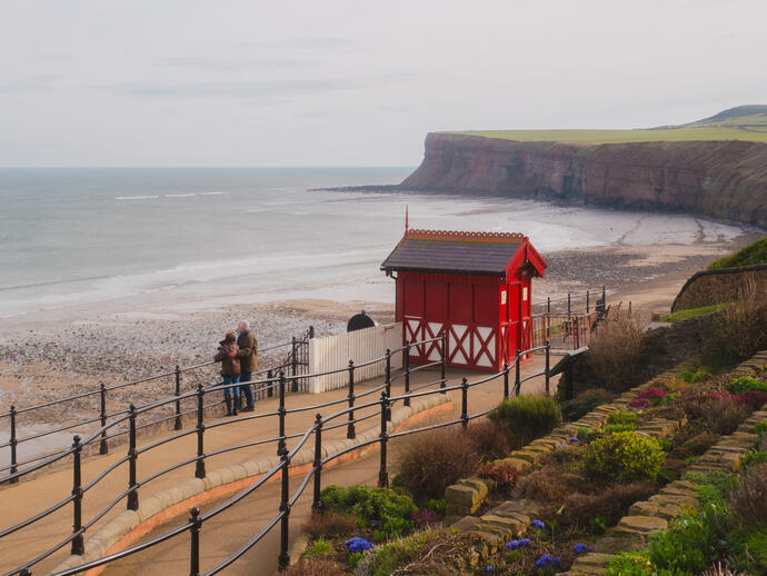 Coastal scene with red beach hut, people on walkway, cliffs, and gardens overlooking a sandy bay.
