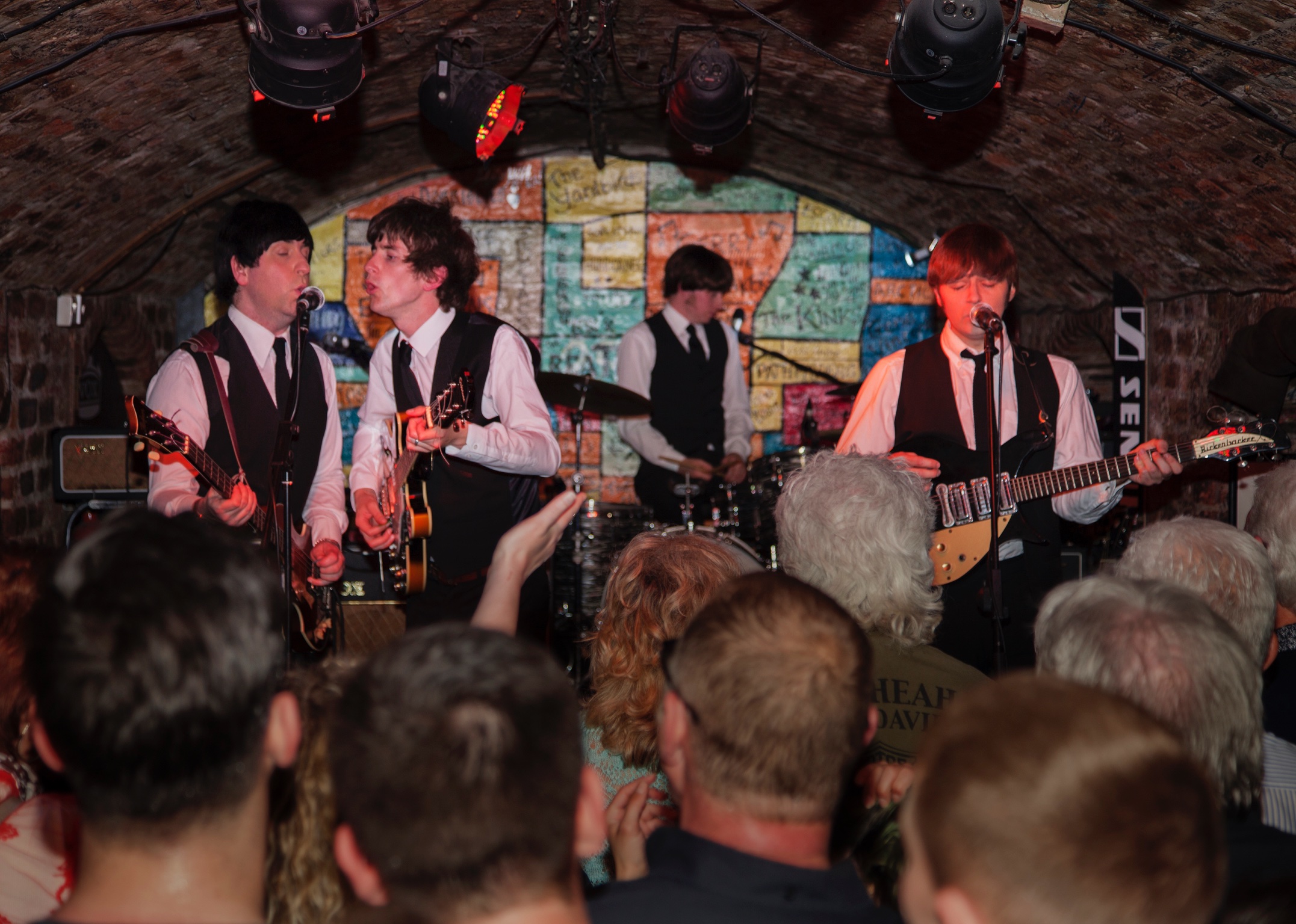 A band dressed in matching black vests and ties performs live in a crowded, arched brick venue with a colorful graffiti-style wall in the background.