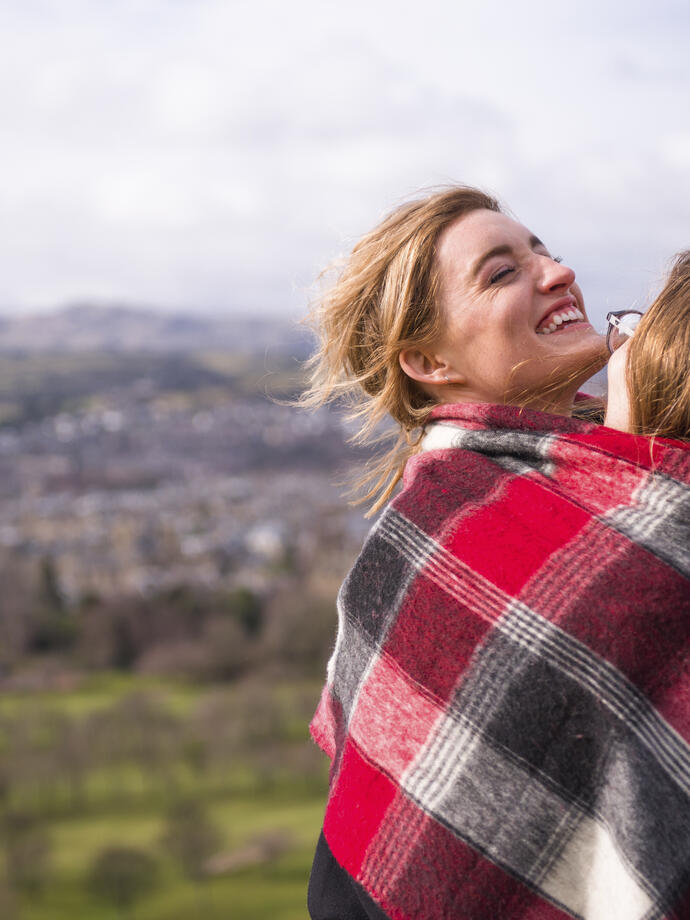 Two women wrapped in red checkered blanket standing on a hill overlooking a city
