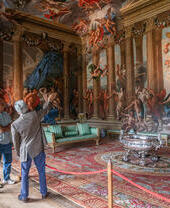 A man and a woman with a tour guide in an ornate room with paintings
