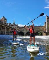 Two people paddleboarding down a river in the centre of Bath