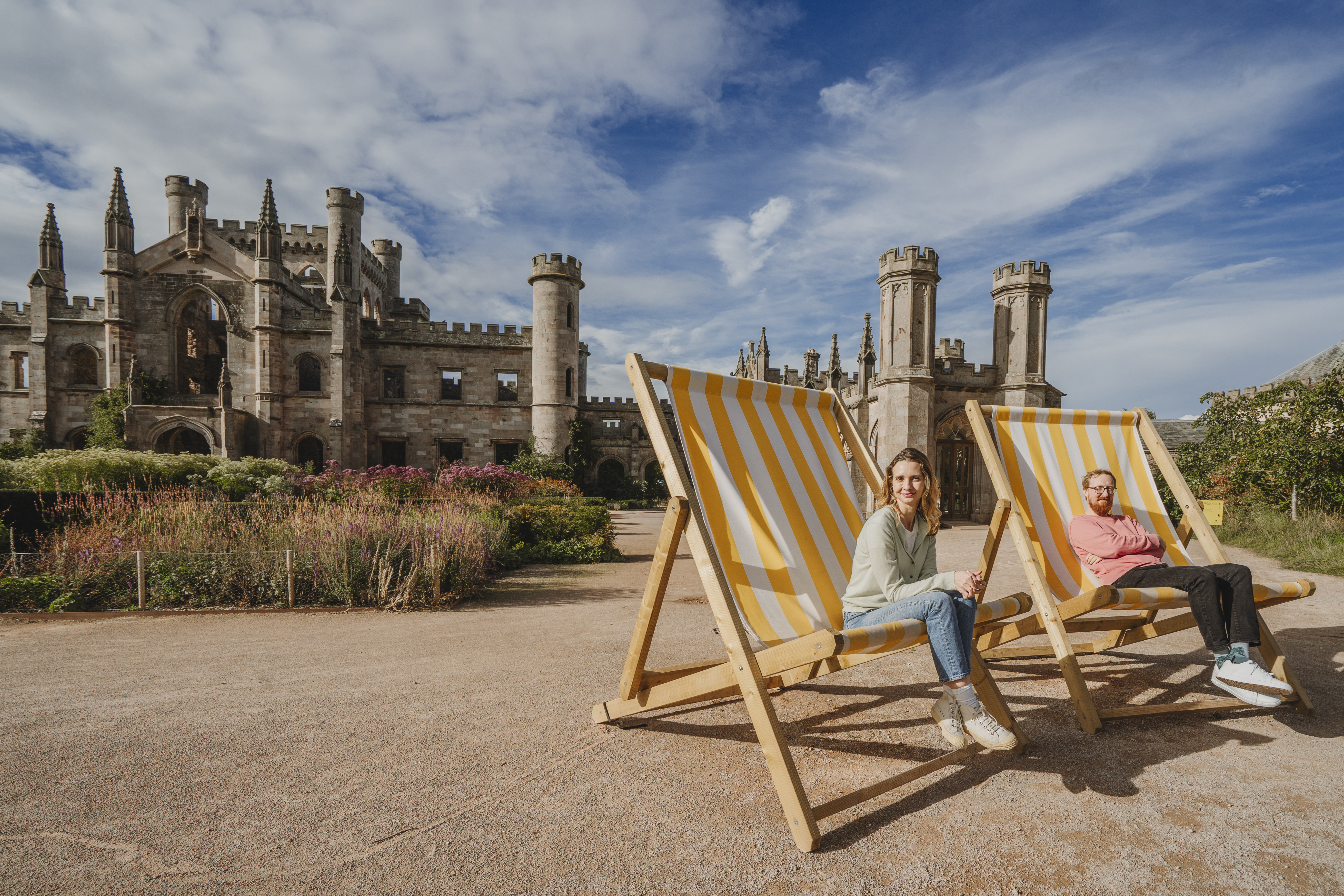 Deux personnes assises sur d’énormes chaises longues rayées jaune et blanc devant un grand château historique avec tours et jardins sous un ciel bleu.